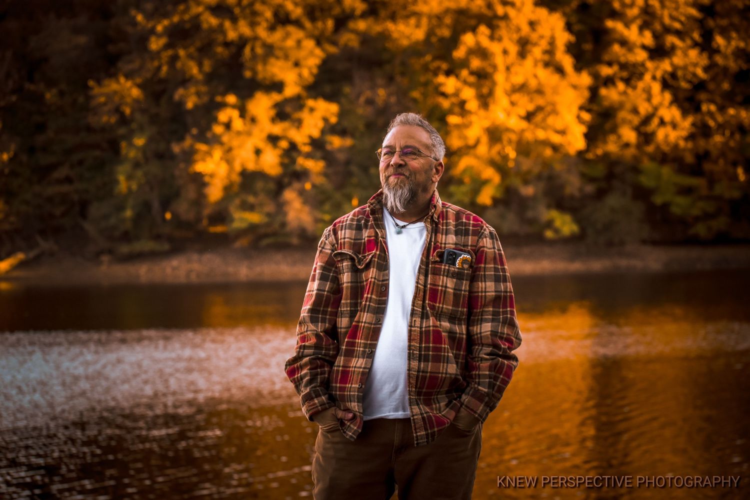 Person in plaid flannel shirt stands against vibrant autumn foliage at sunset by a serene lake.