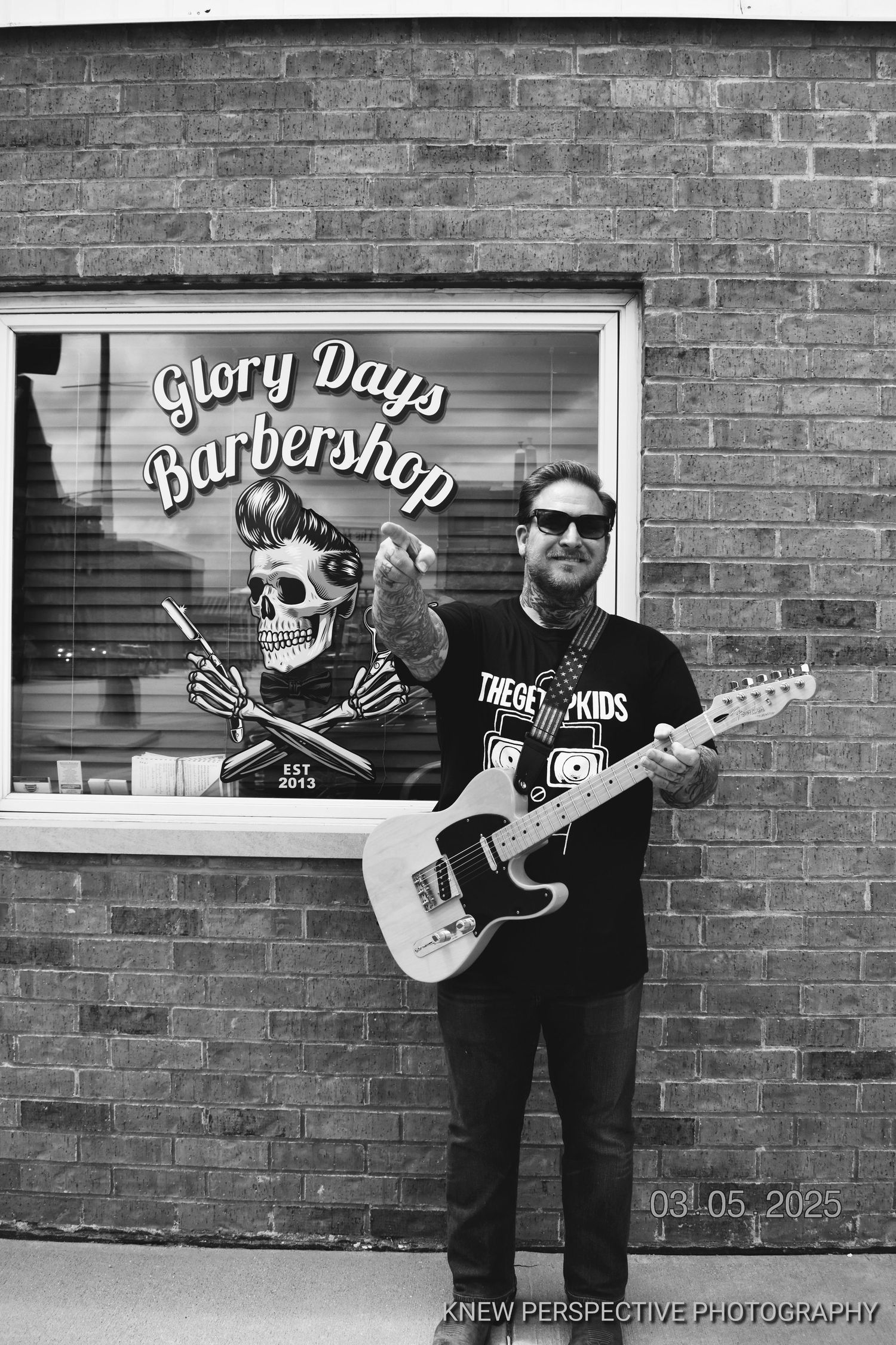 Black and white photo showing business owner Kyle Rohweder outside Glory Days Barbershop with rockabilly skull artwork in the window.