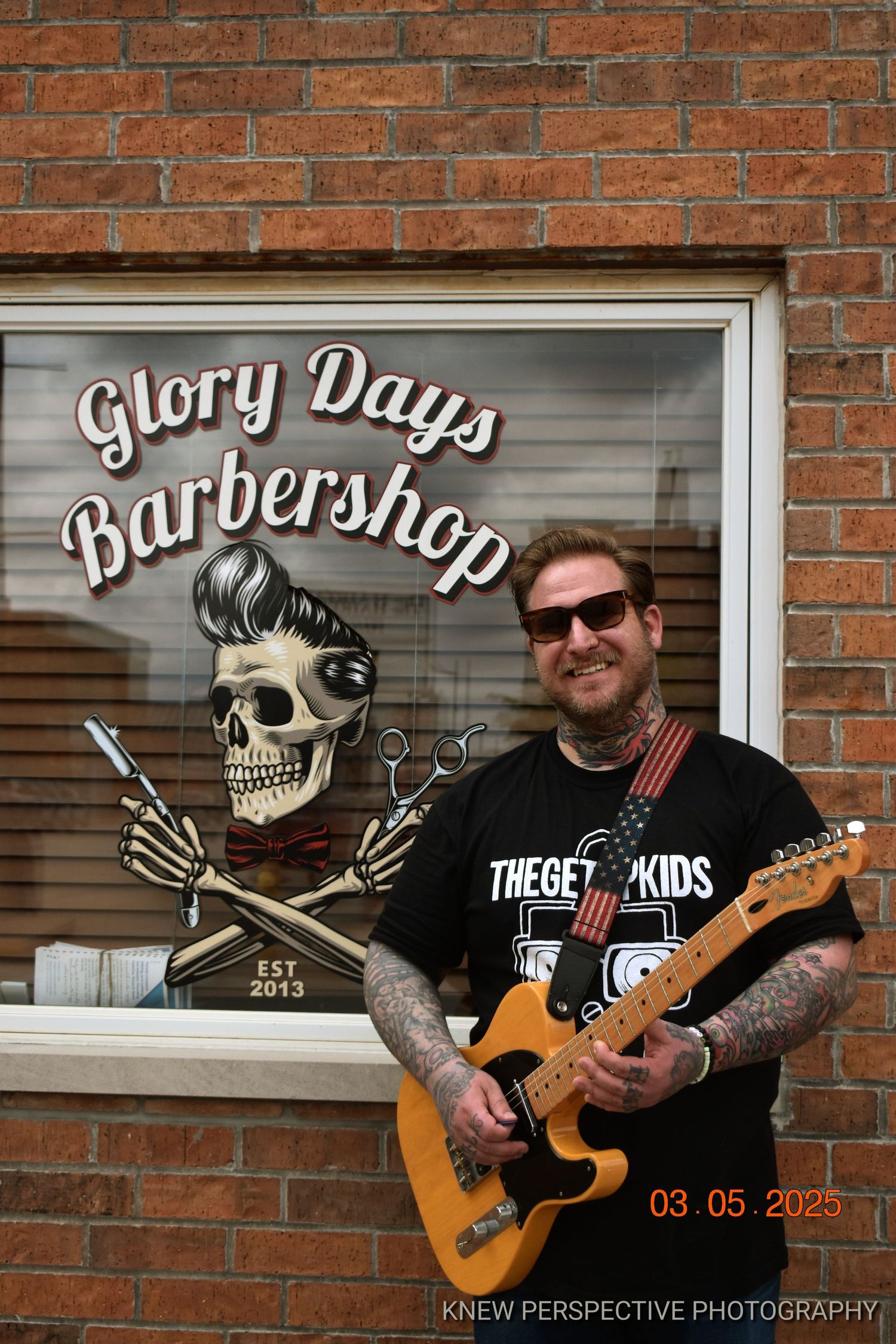 Barber shop storefront with skull logo and guitar player sitting outside brick building with vintage signage.