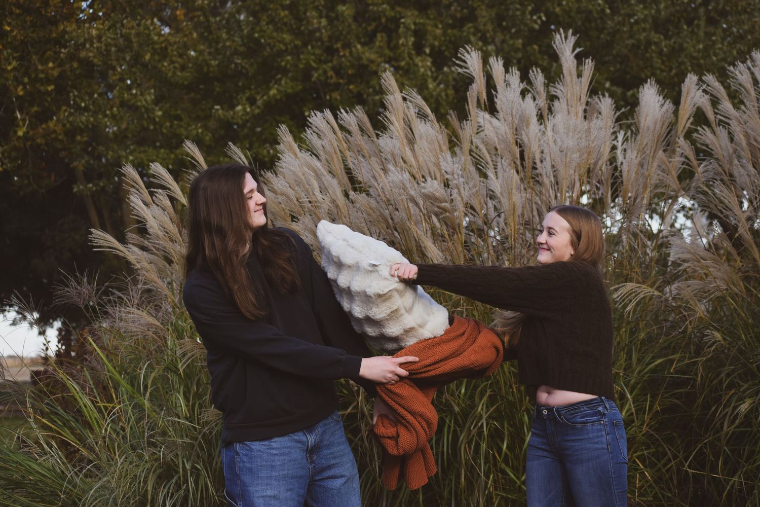 Two people playfully tug at a blanket while standing in front of tall ornamental pampas grass on an autumn day.