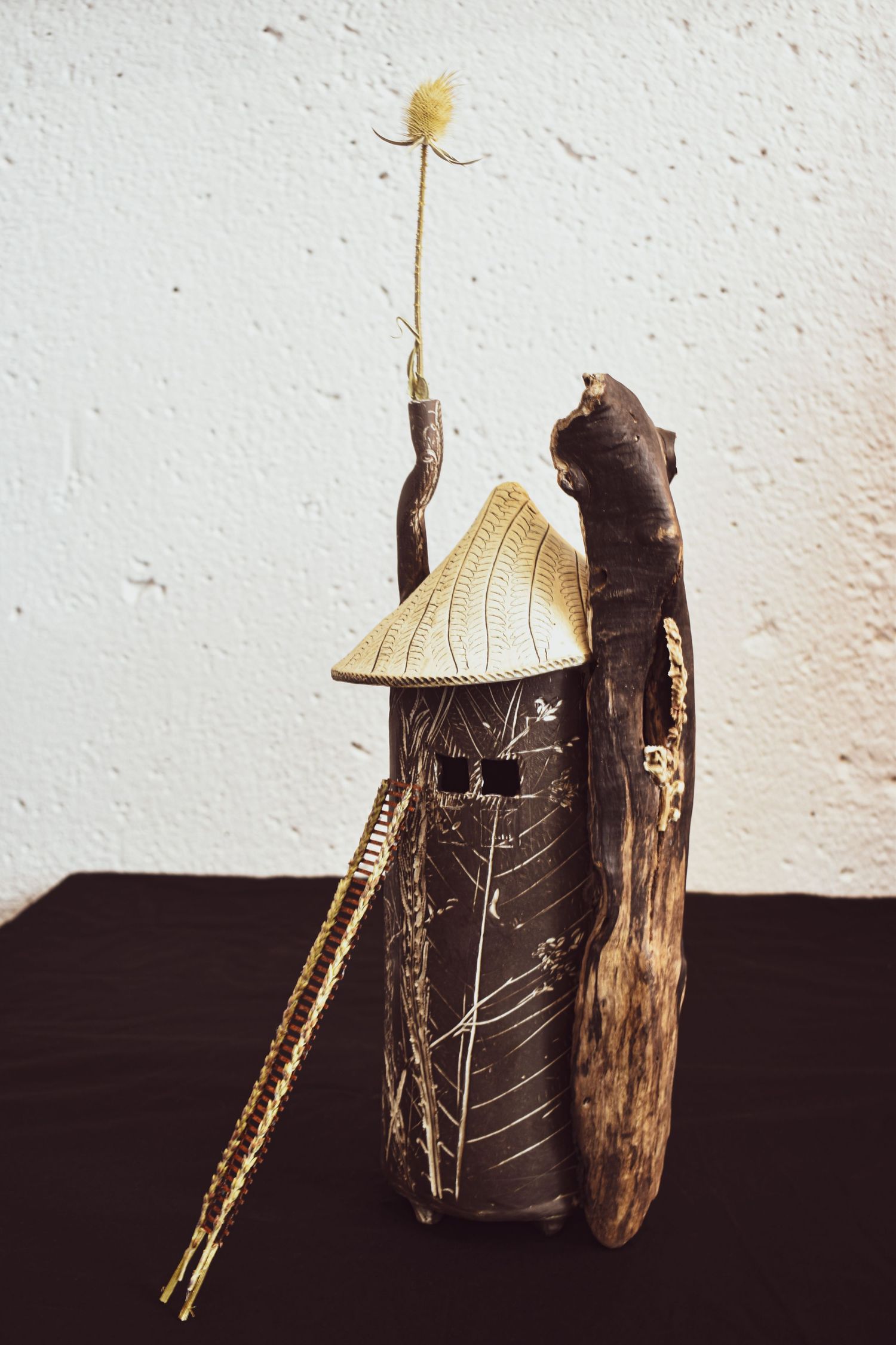 Artistic sculpture made from clay and natural materials featuring a conical hat roof and delicate flower on top.