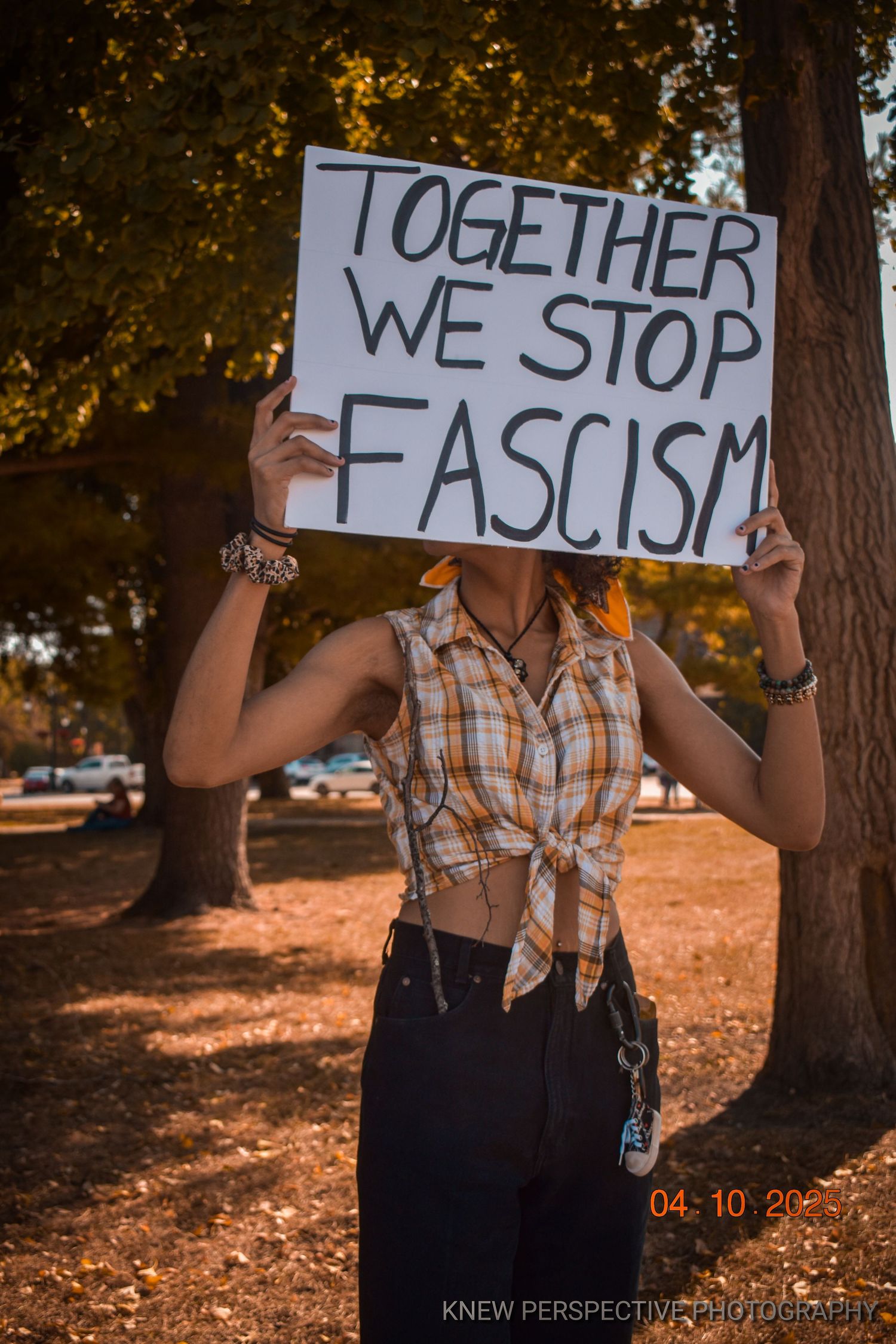 A protest sign reading 'Together We Stop Fascism' held up against a backdrop of autumn trees at sunset.