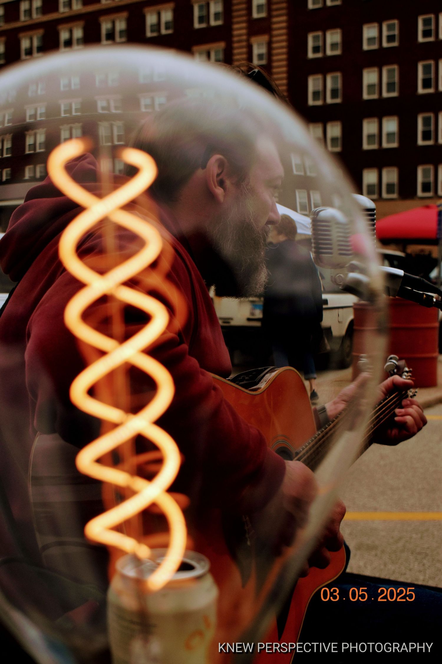 Artistic photo of a musician playing guitar with glowing orange neon spiral light against urban building backdrop.