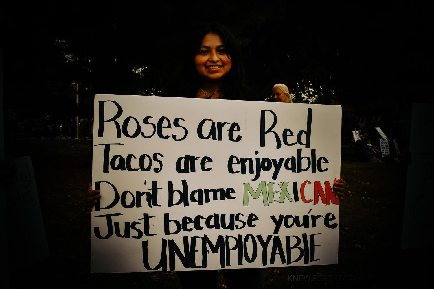 A Latino woman holding a protest sign that reads 'roses are red tacos are enjoyable don't blame Mexicans just because you're unemployable'