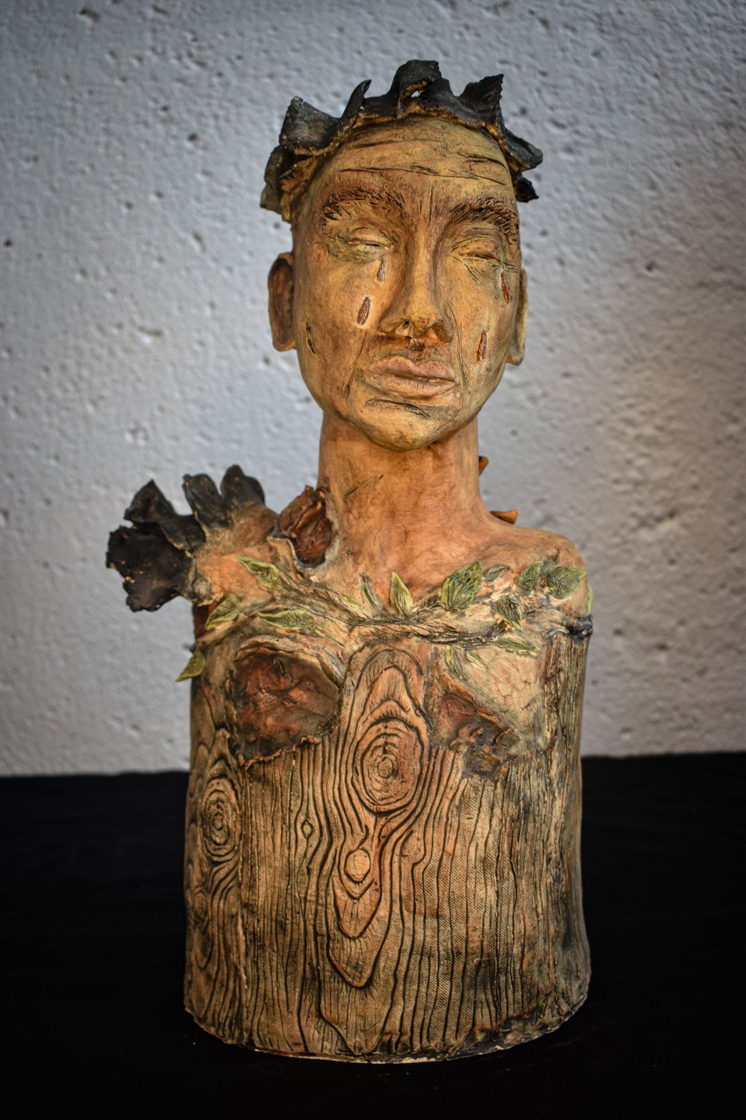 A handcrafted ceramic bust with wood-like texture and leaf details shows a contemplative expression against a white wall.
