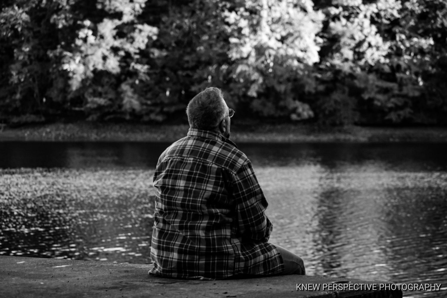 A solitary figure in a plaid shirt sits contemplatively by a tranquil Lake Storey on a peaceful afternoon.