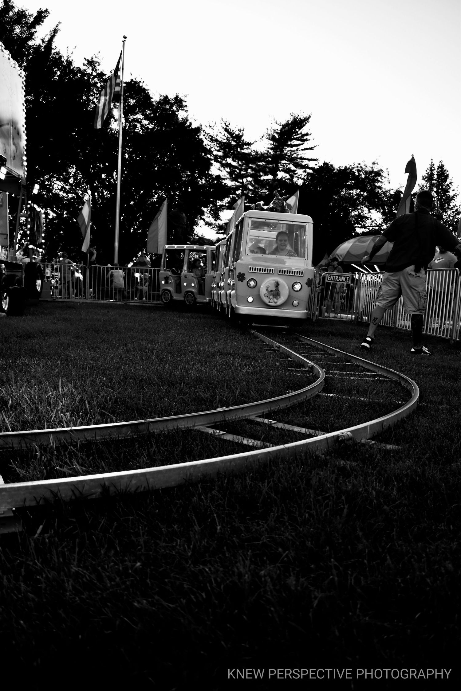Black and white photo of a miniature train ride on curved tracks at a carnival at dusk.