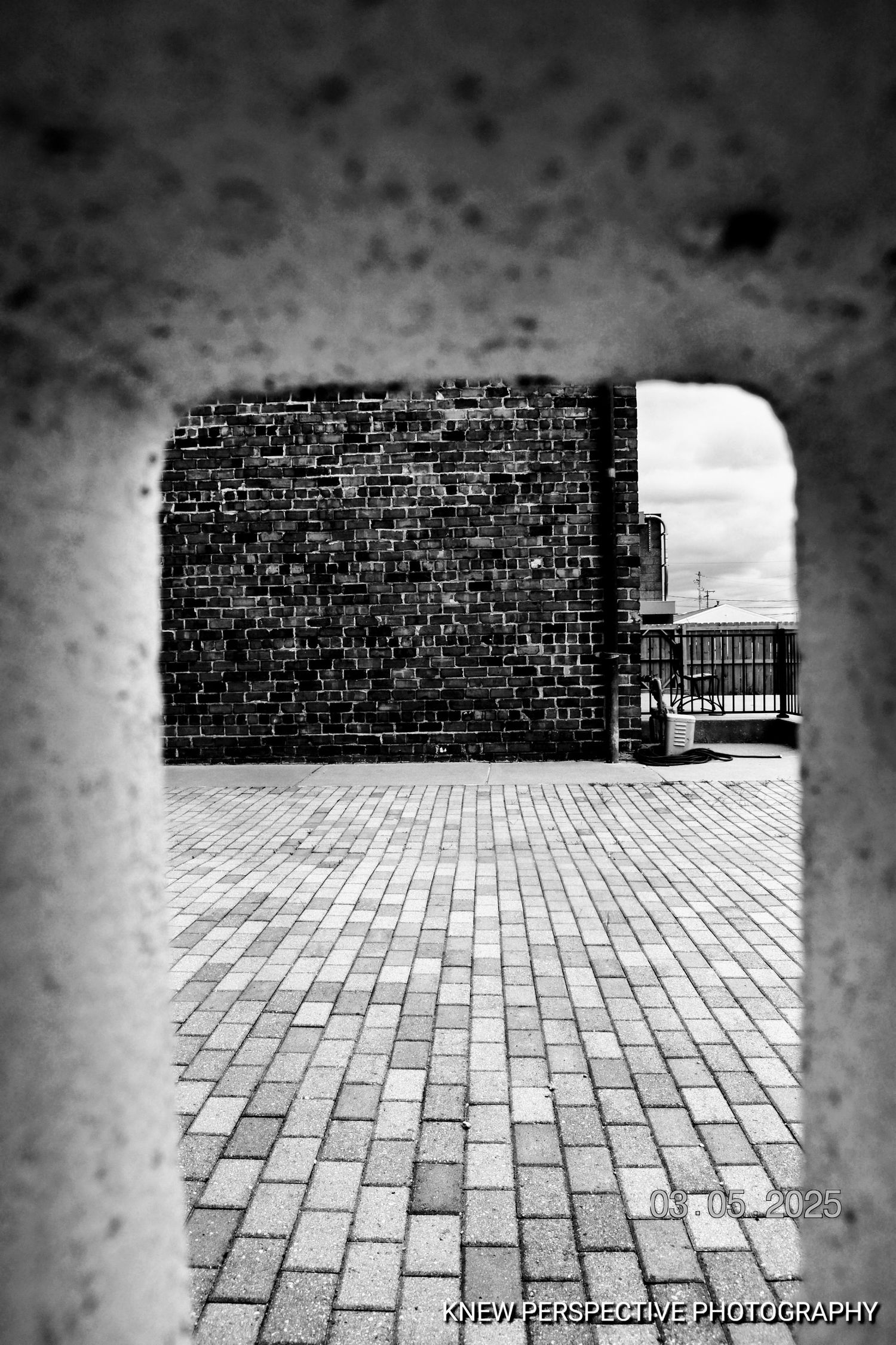 Black and white architectural photograph showing brick wall and paved courtyard through cinder block opening.