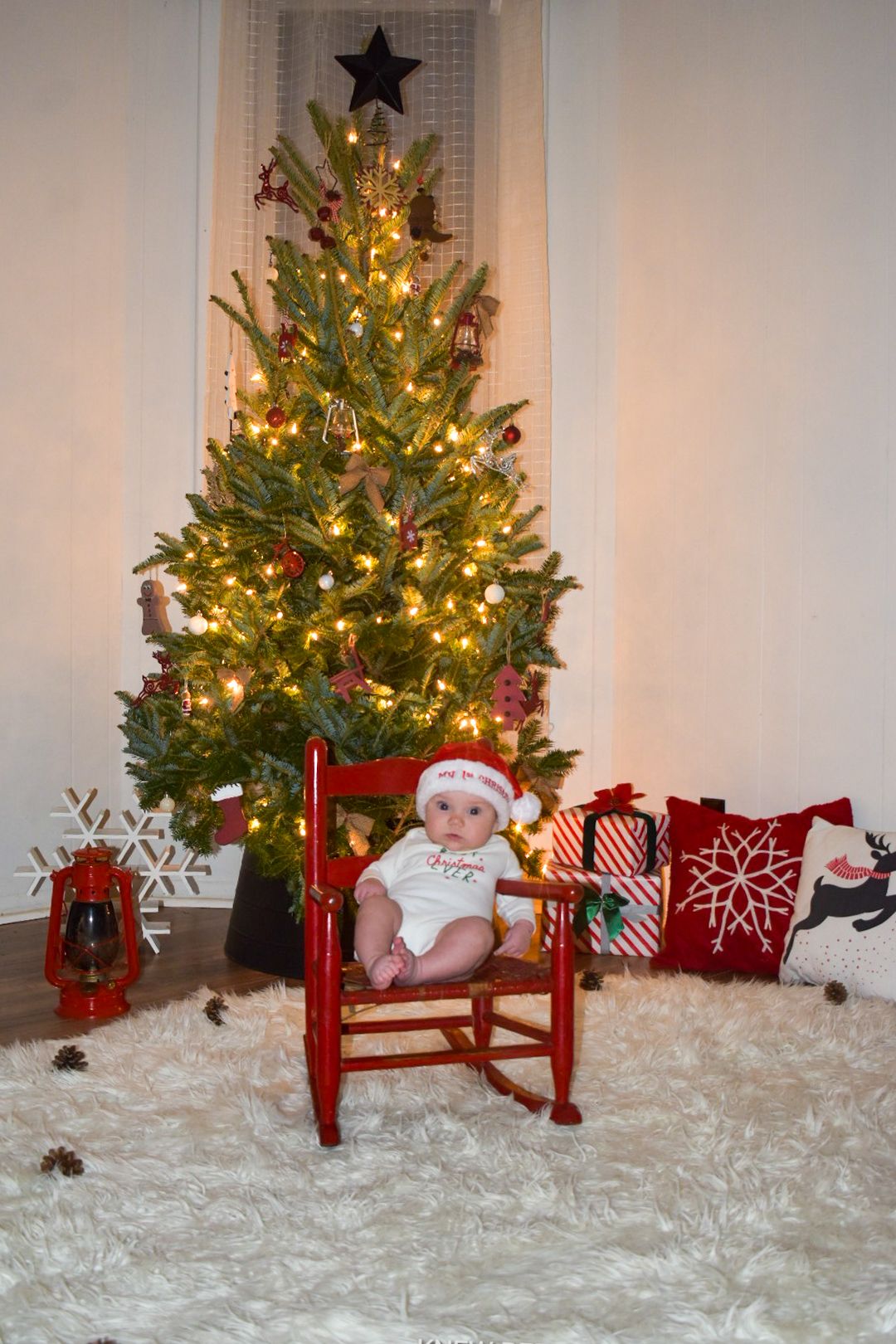 Decorated Christmas tree with twinkling lights next to a baby nestled in a little red rocking chair, on white carpet.
