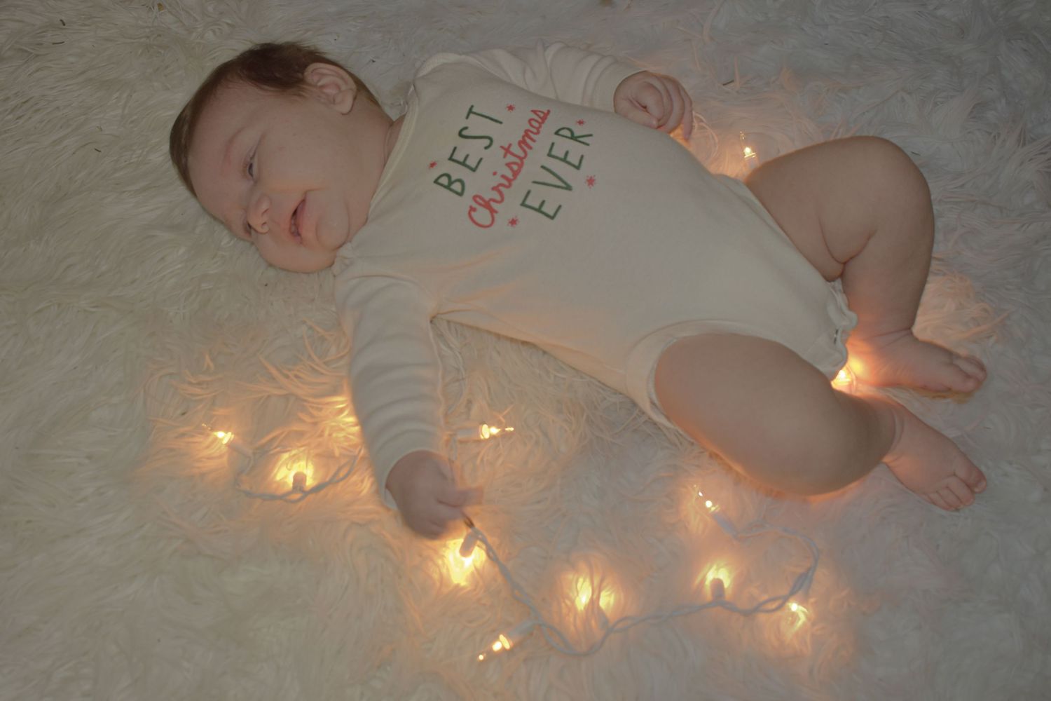 A sleeping baby in white onesie lies on soft fabric surrounded by twinkling fairy lights.