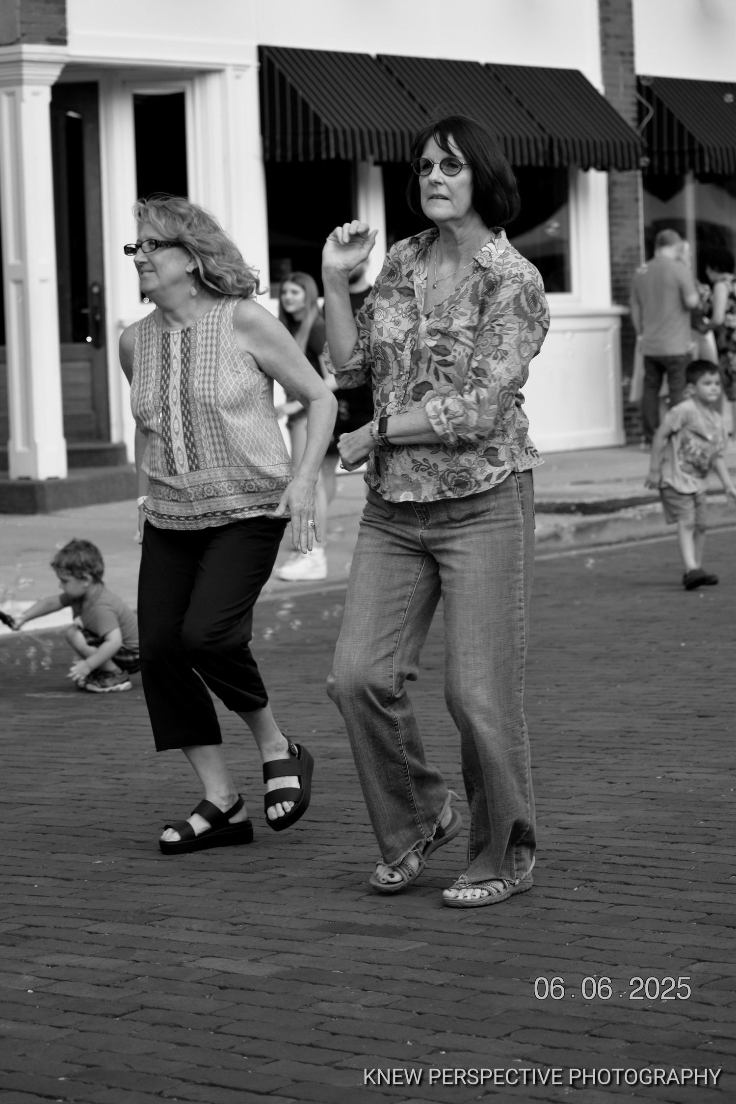 Black and white photograph showing feet and legs of people dancing on a brick road in a casual outdoor setting.