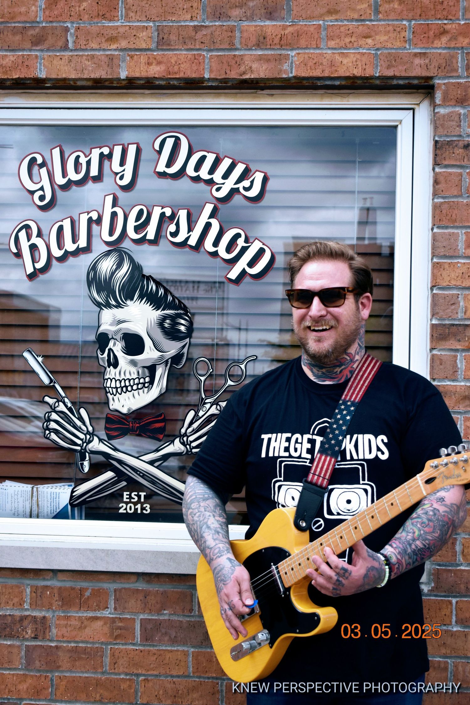 Guitarist performs in front of brick wall with barbershop skull logo and signage visible in background.