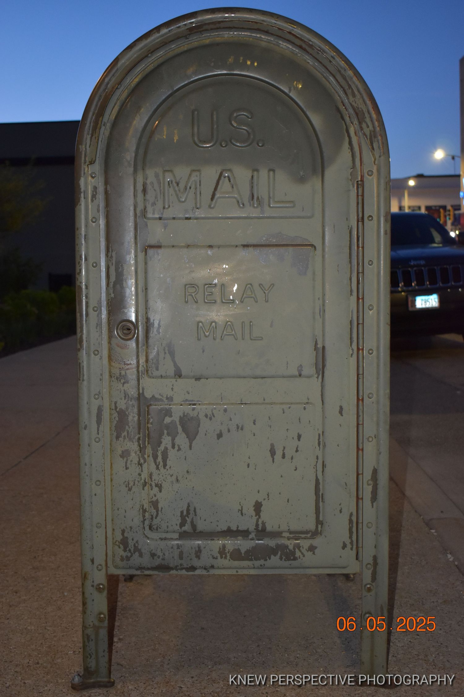 An old weathered U.S. Mail relay box stands against a dark background with a moon visible in the evening sky.