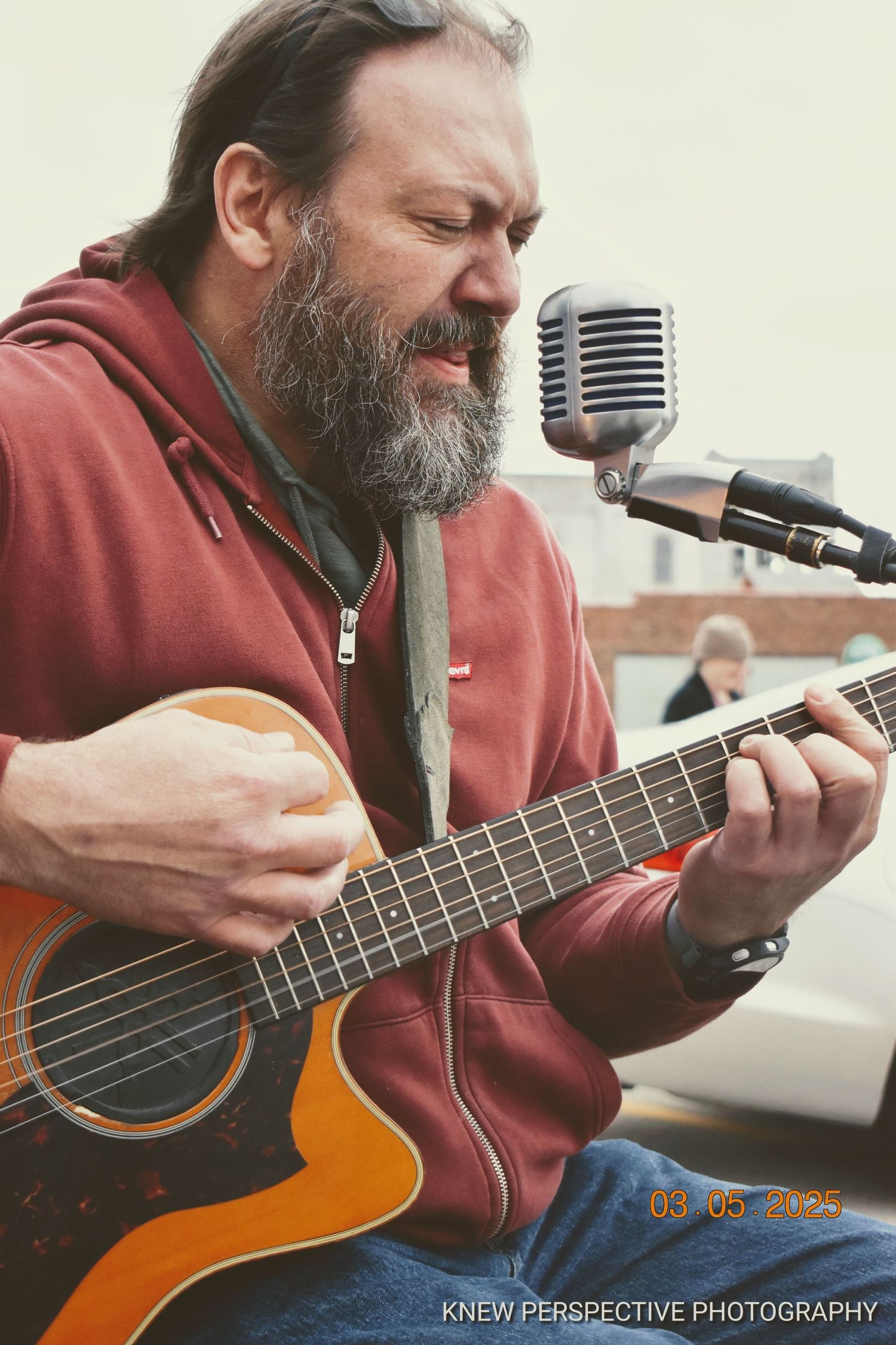 Musician in red hoodie plays acoustic guitar and sings into vintage microphone during outdoor performance.
