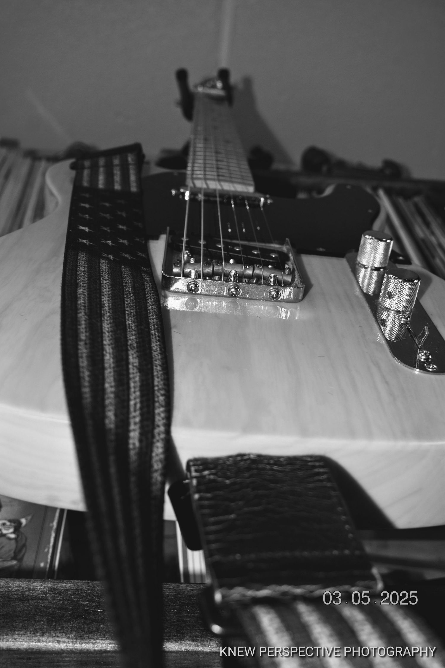 Black and white close-up photograph of an electric guitar showing strings, bridge and controls on a light surface.
