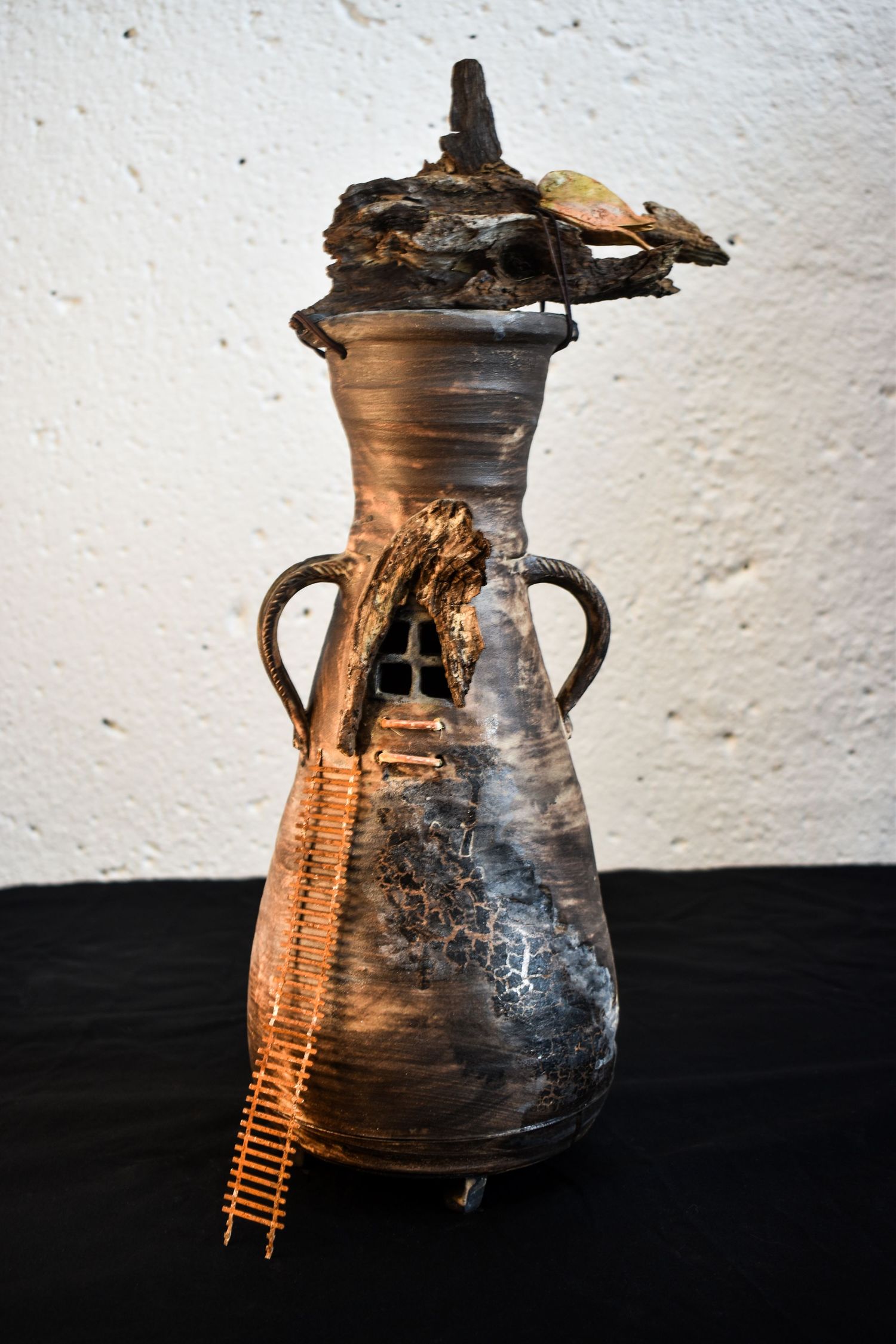 An antique ceramic pot with dark patina and handles photographed against a white wall in dramatic lighting.