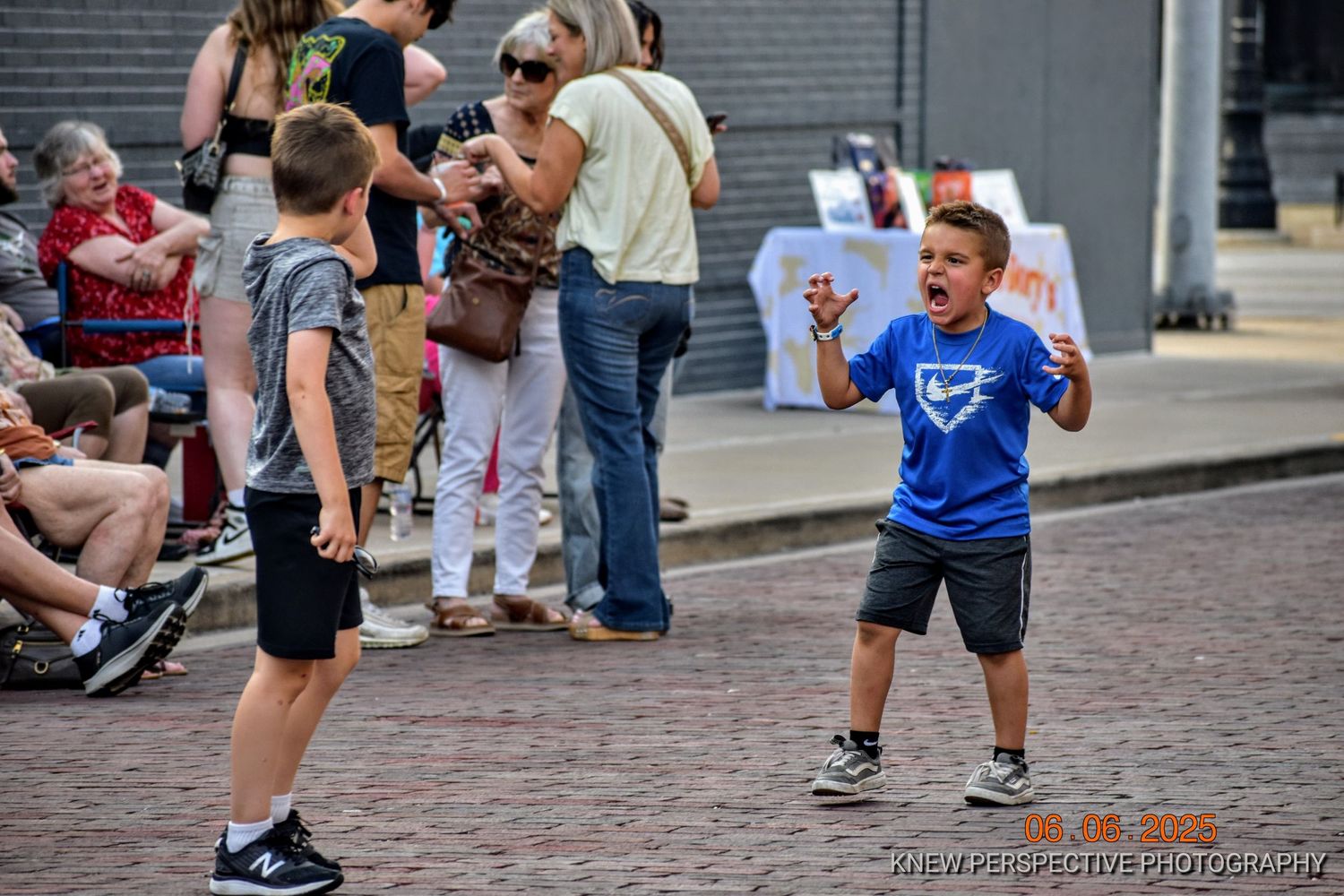 Two children interact during an outdoor event on a brick paved street with people gathering in the background.