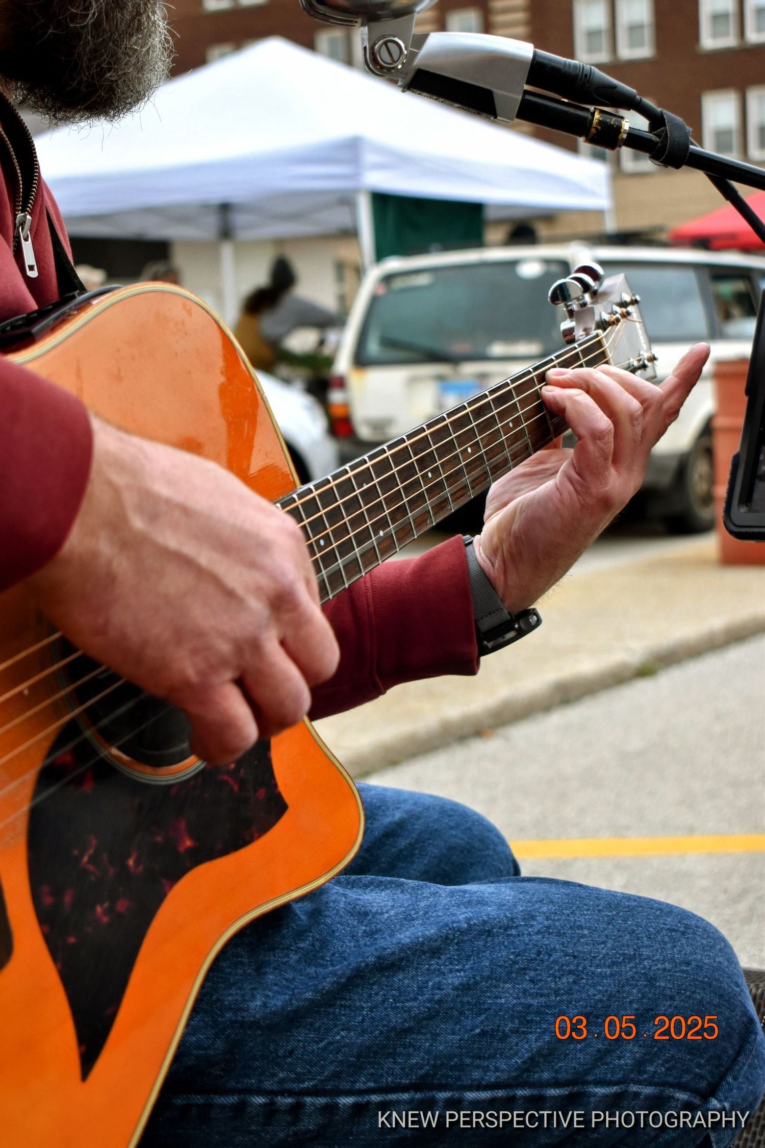 Close-up sequence of hands playing an acoustic guitar at an outdoor event with buildings in the background.