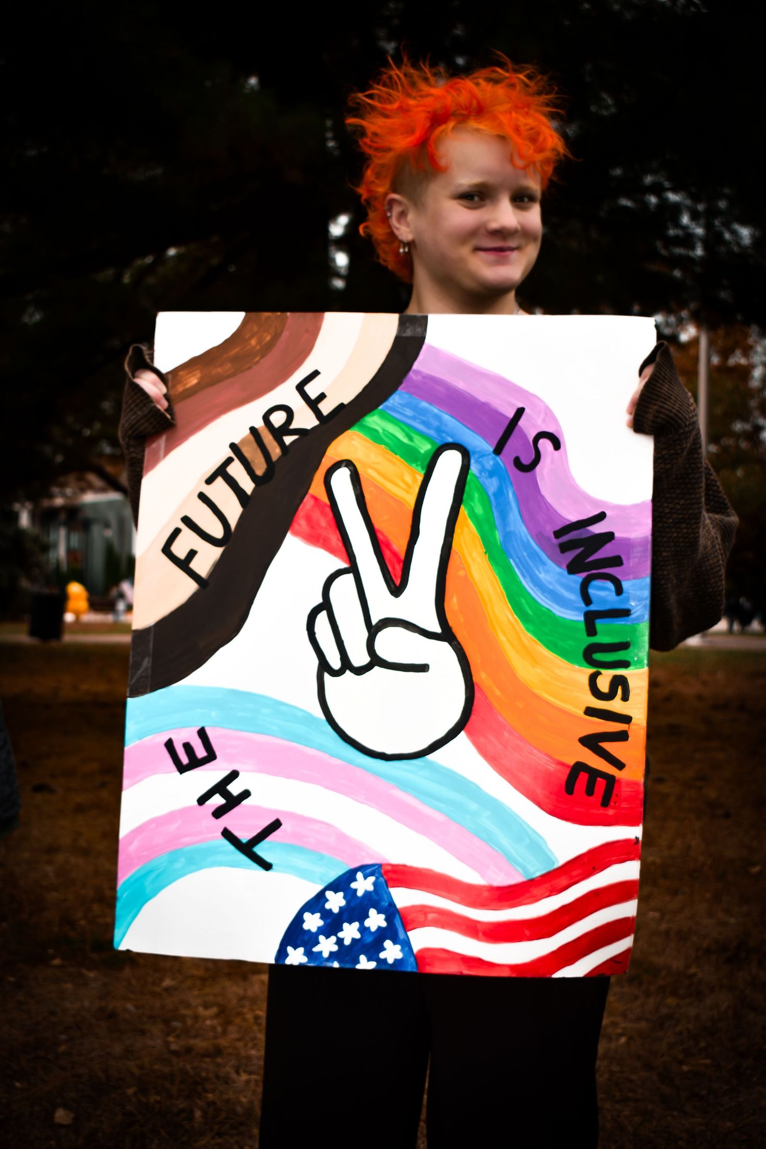 A protest sign featuring rainbow colors, peace symbols, and American flag elements arranged in a progressive sequence.