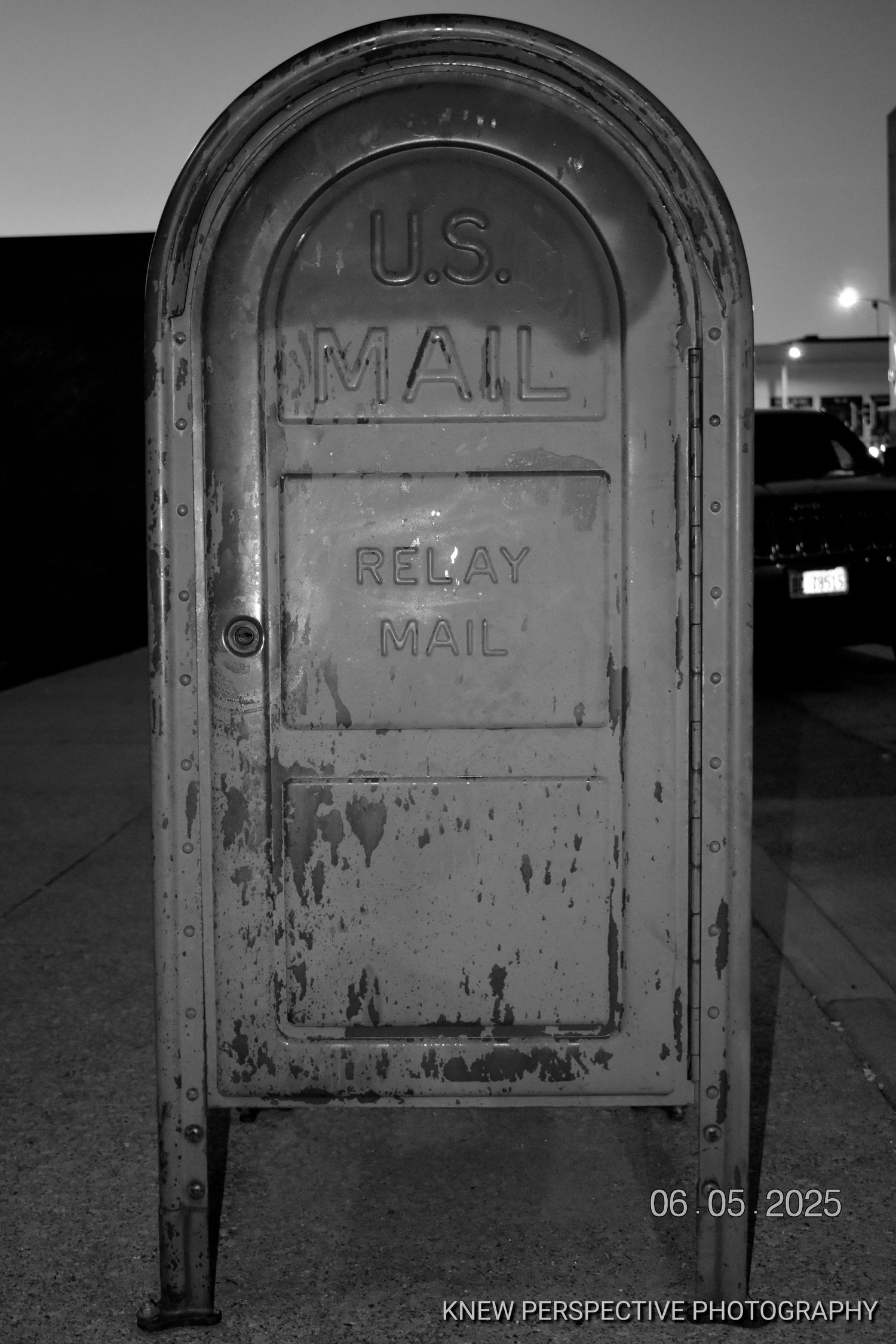 A weathered vintage U.S. Mail relay mailbox photographed in black and white at night with a dim light in the background.
