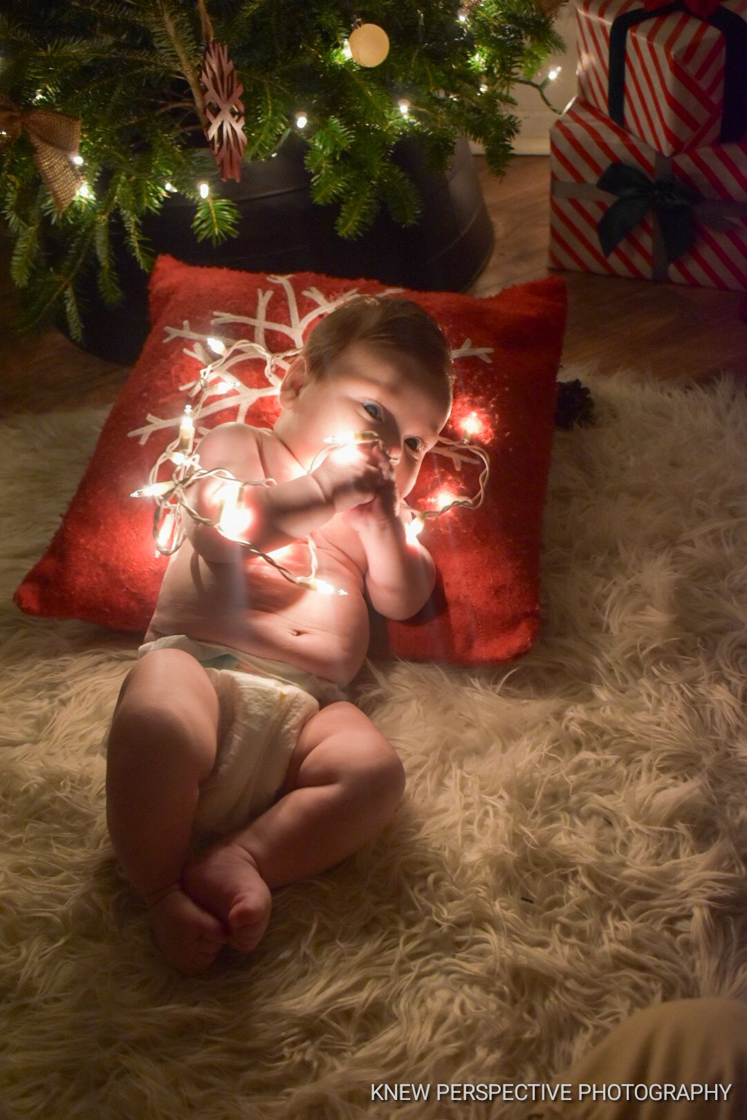 Baby lying on soft fur rug with glowing string lights under Christmas tree decorations creating magical holiday scene.