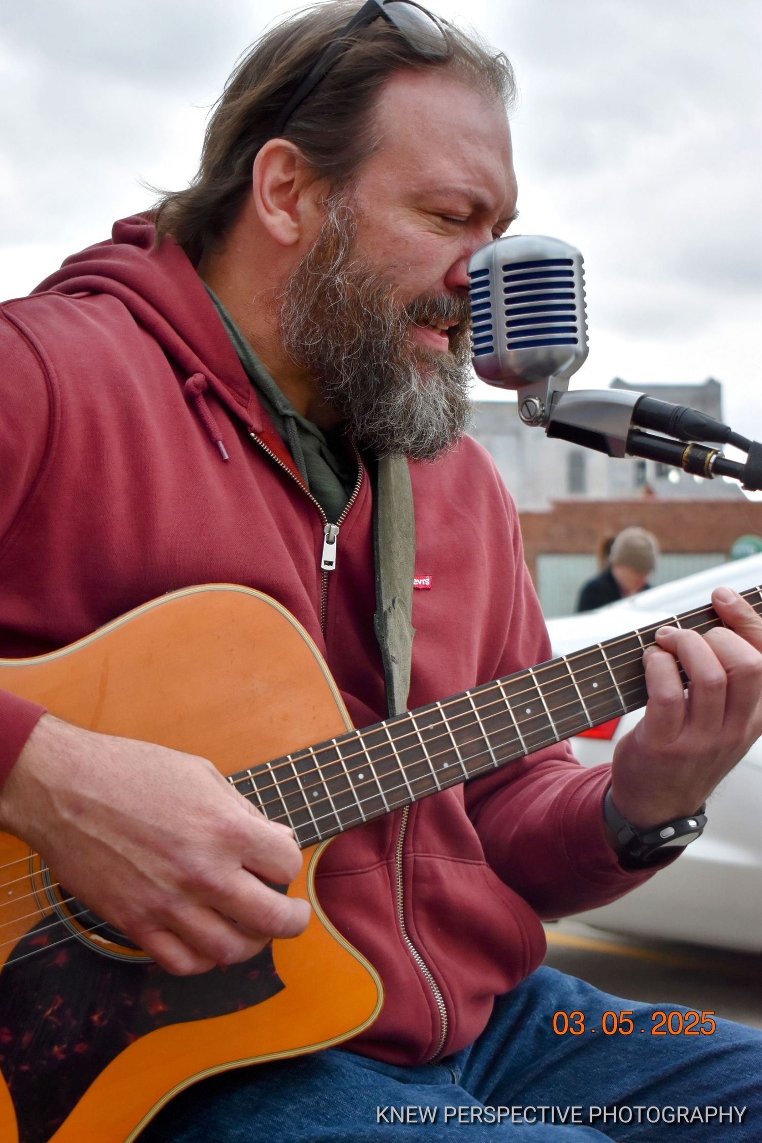 Musician in burgundy hoodie performs with acoustic guitar and microphone at outdoor venue.