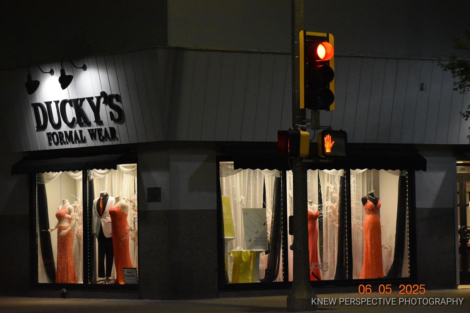 A dimly lit storefront at night with glowing window displays and illuminated signage reading 'Ducky's Formal Wear'.