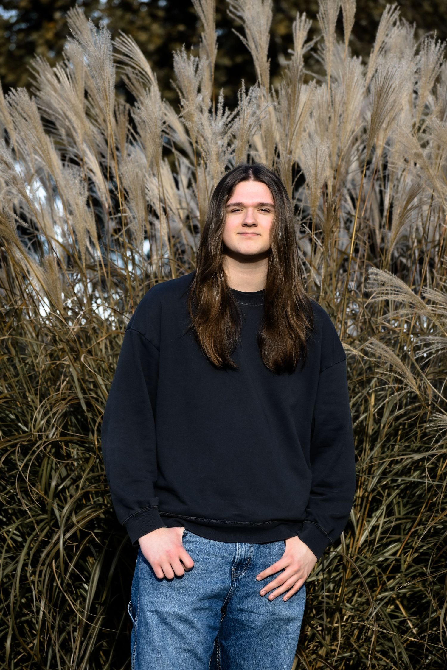 A person in a black sweater and jeans stands in front of tall pampas grass on a sunny day.