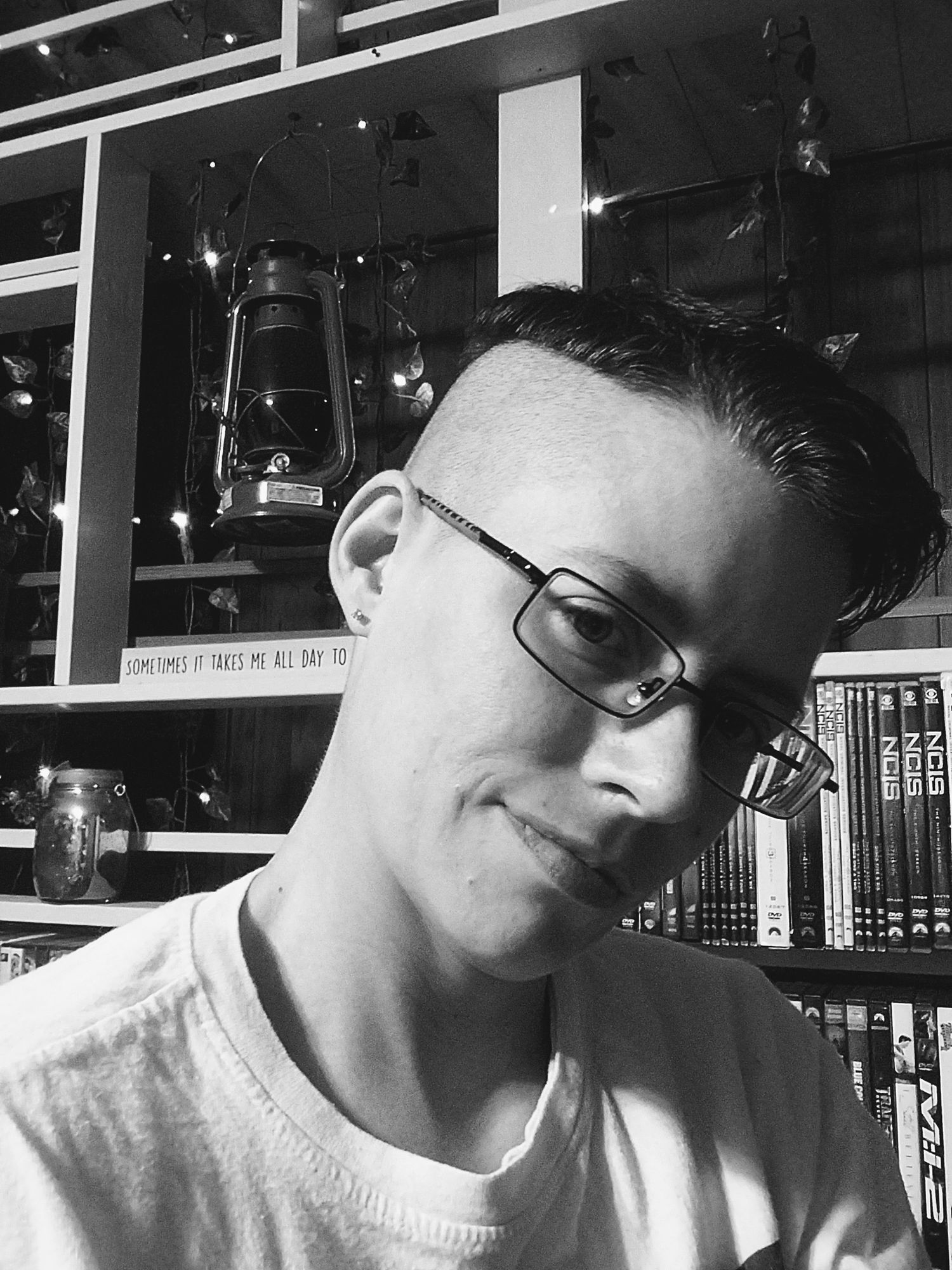 A black and white photo shows someone wearing glasses against a background of bookshelves and lanterns.