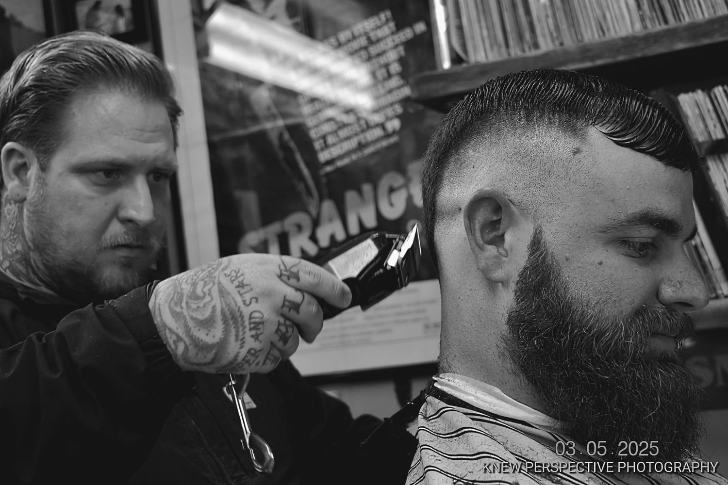 A barber with tattoos works on a client's haircut in a black and white photo at a barbershop.