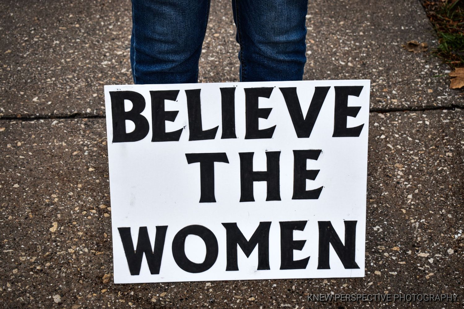 A protest sign spelling 'BELIEVE THE WOMEN' in bold black letters.