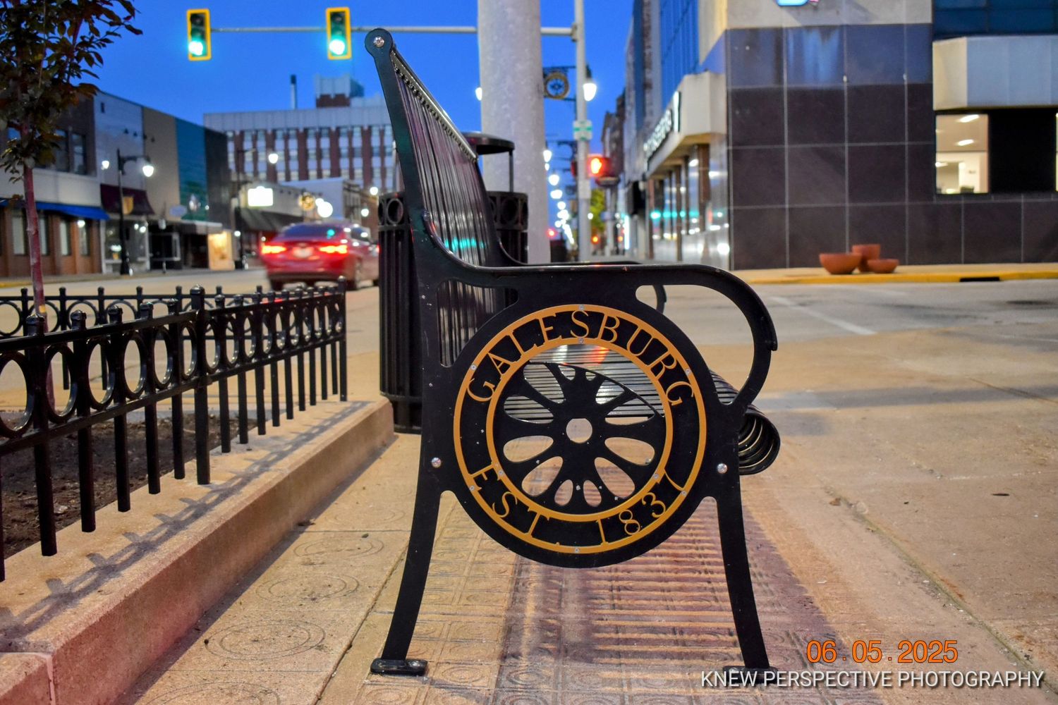 Decorative metal bench with wheel design sits along a city sidewalk with black railing and traffic lights in background.