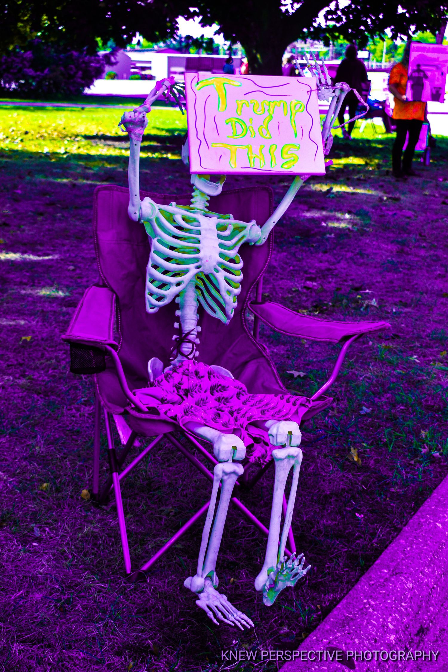 A skeleton decoration sits in a purple camping chair holding a sign under neon-colored lighting in an outdoor setting.