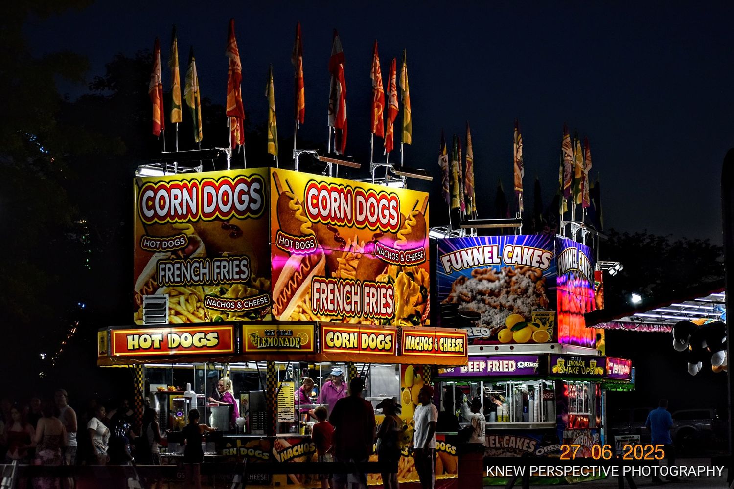 Illuminated carnival food vendor stands selling corn dogs and snacks with bright neon signs at night.