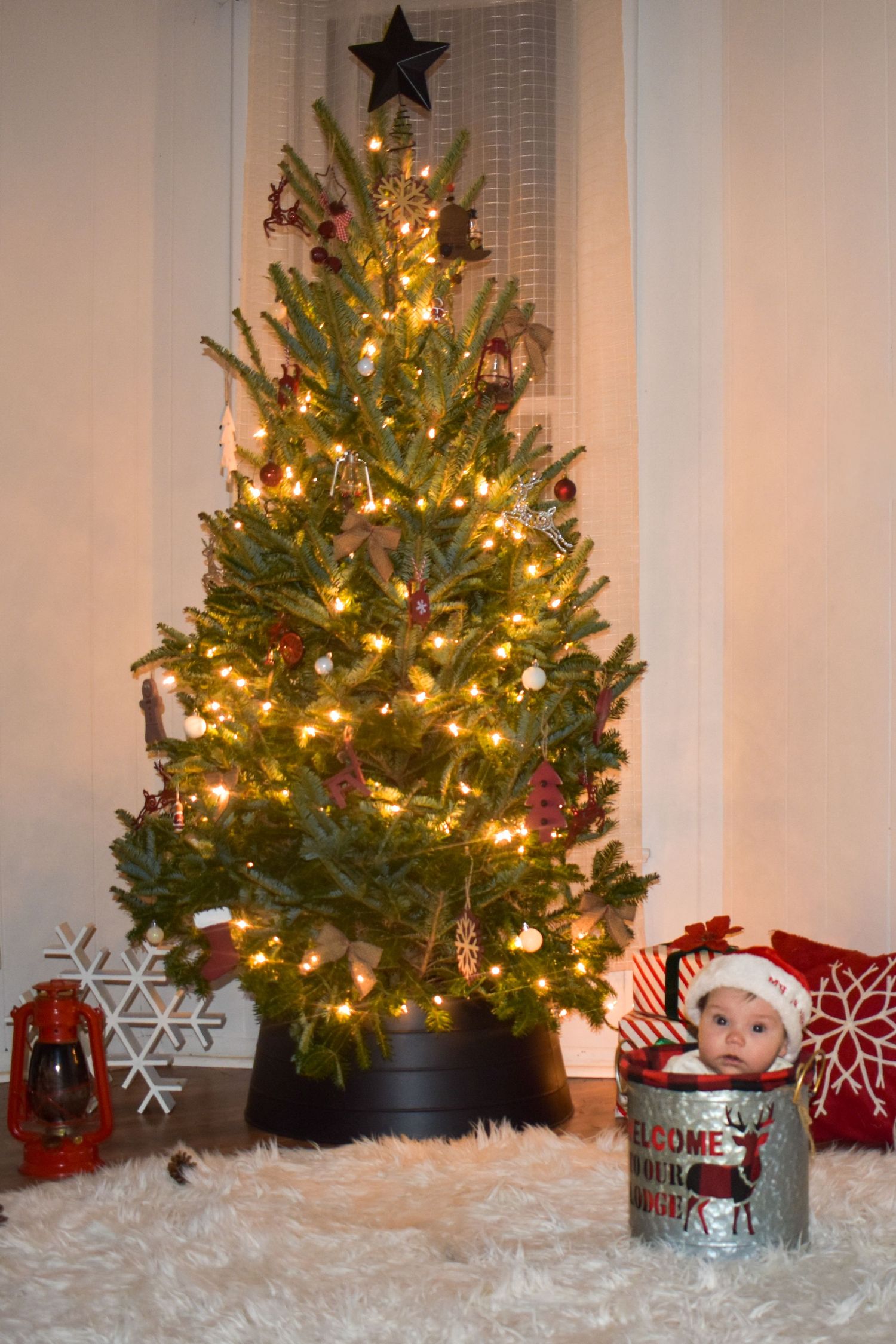 Illuminated Christmas tree behind a baby sitting in a  galvanized bucket.