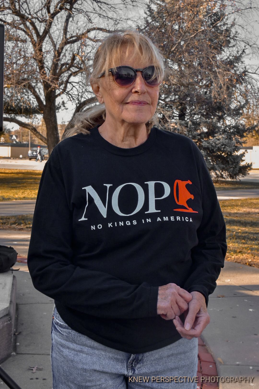 Person wearing sunglasses and a black sweatshirt with 'NOPE No Kings in America' text standing outdoors in fall.