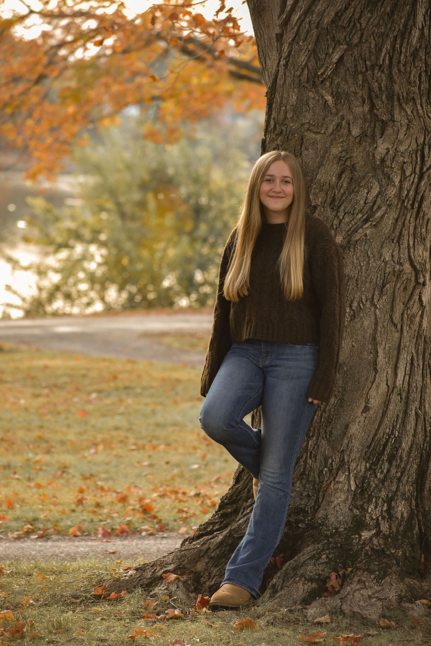 A person in a brown sweater and jeans leans against a large tree trunk surrounded by autumn foliage.