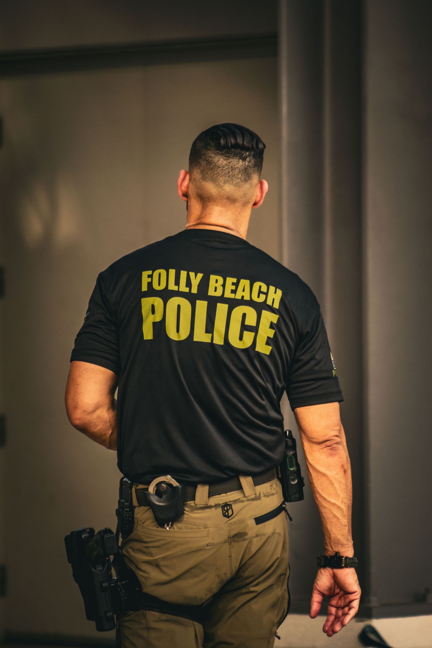 Folly Beach Police officer seen from behind wearing black uniform shirt with gold lettering and duty belt.