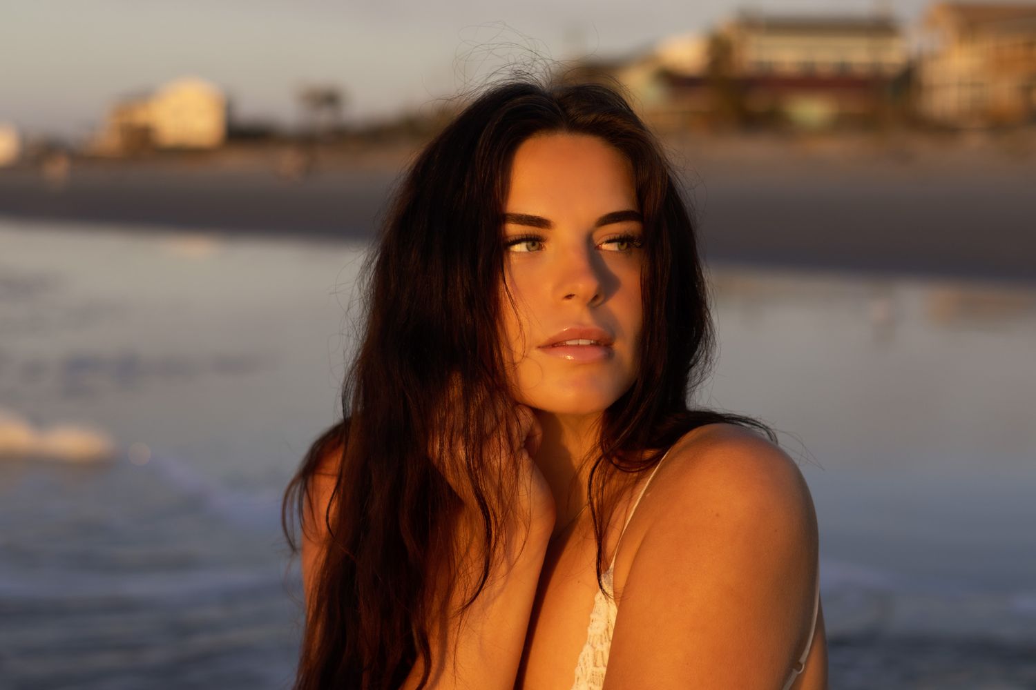 Young woman with long dark hair poses on a sandy beach at golden hour with ocean waves in the background.
