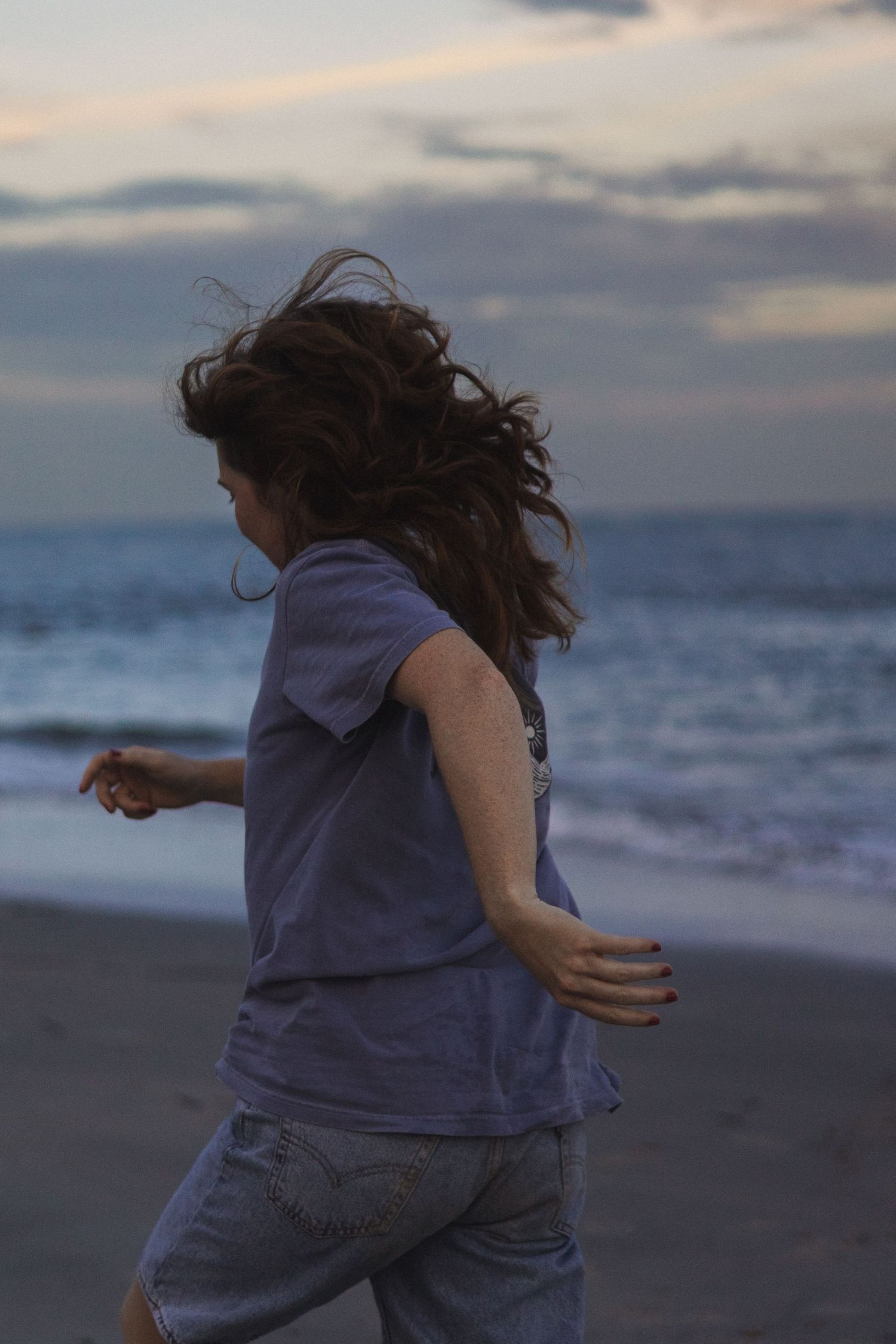Young girl with flowing curly hair running along a beach at dusk with ocean waves in background.