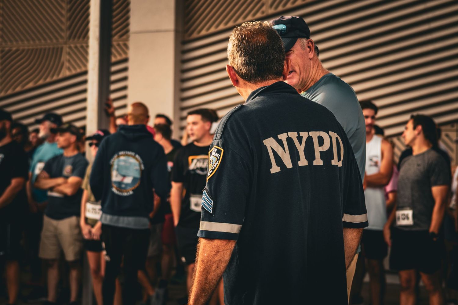 Man wearing NYPD jersey standing among a crowd of people gathered outdoors in warm lighting.