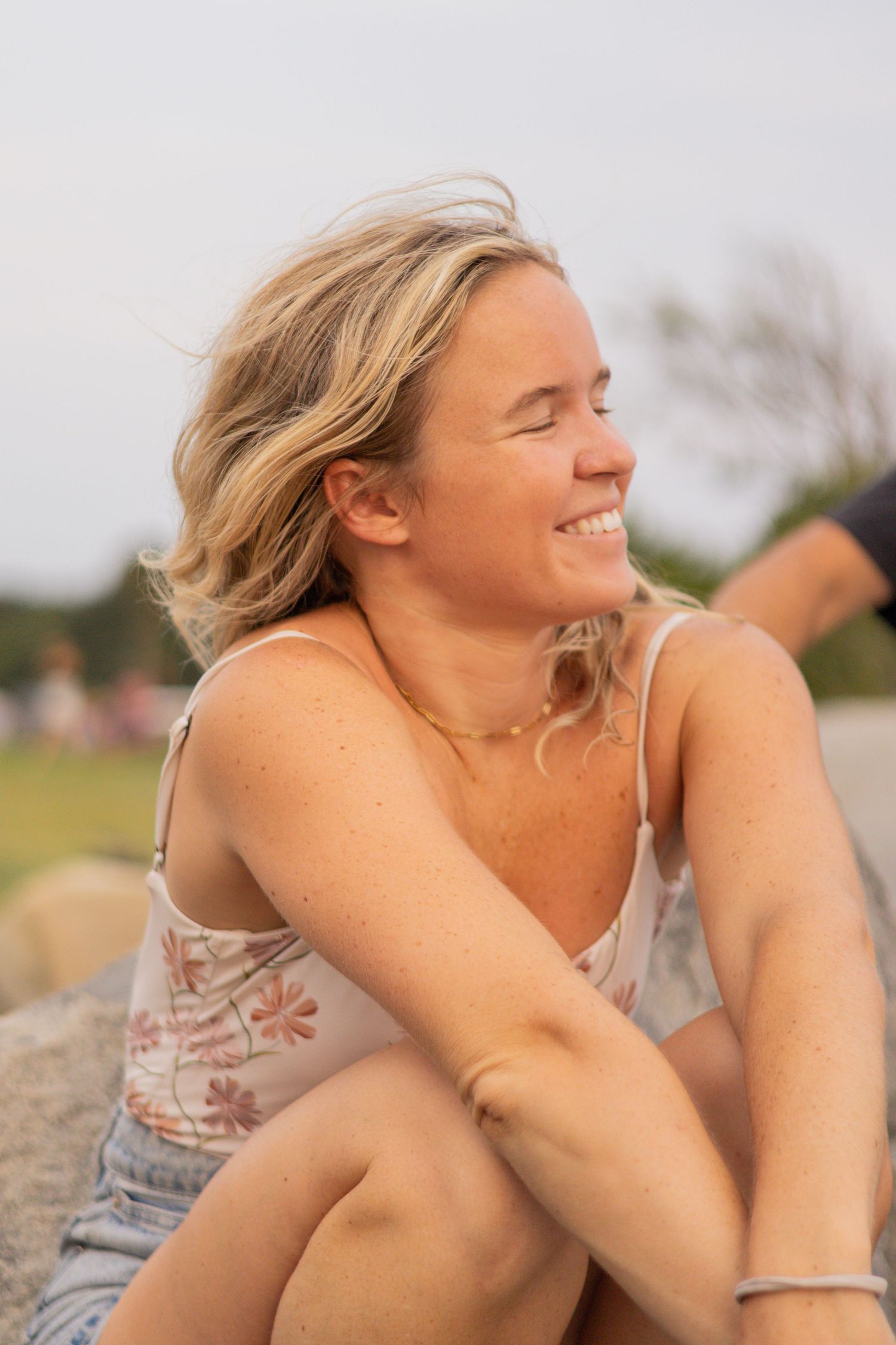 Smiling blonde woman sitting on rocks outdoors, wearing a floral tank top and bracelet, enjoying a sunny day.