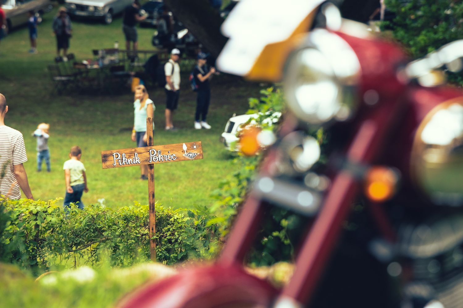 Autós piknik hangulatos nyüzsgése. Piknik Pokróc tábla mutatja, hol találhatóak a plédek. Előtérben egy motor részlete van.