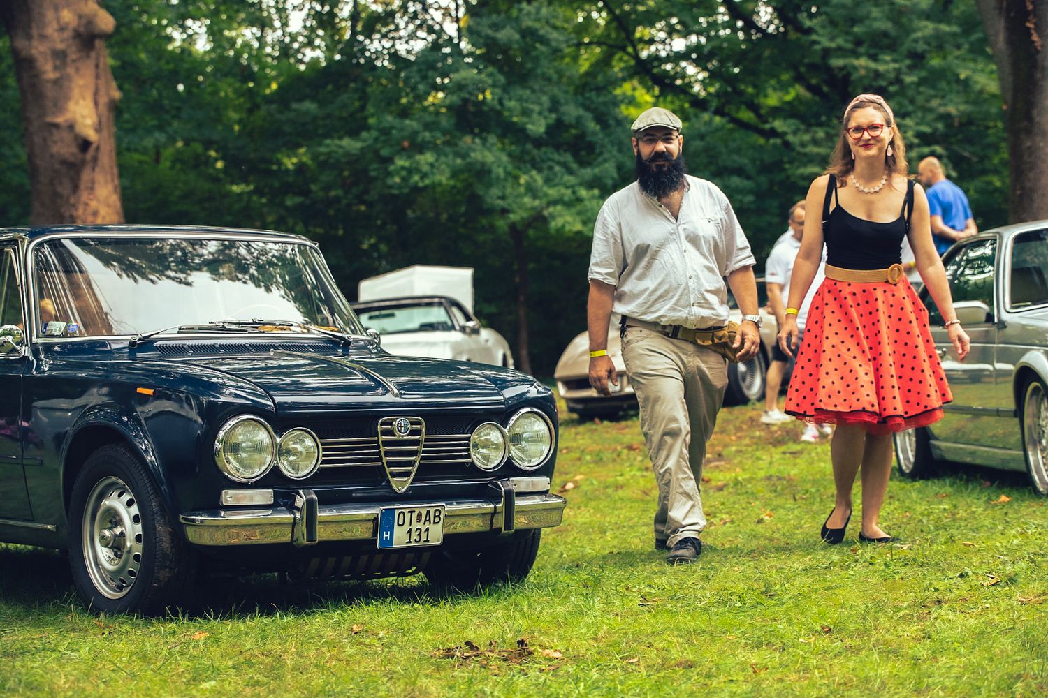 Egy kedves pár sétál a kamera felé egy old timer Alfa Romeo mellett.