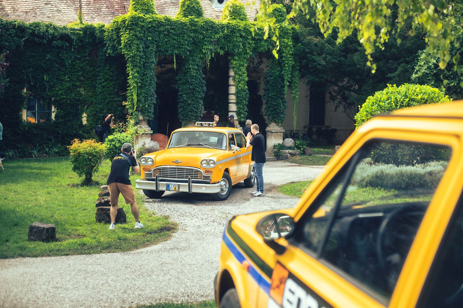 Autó fotózás zajlik egy karakteres sárga old timer taxival egy borostyánnal benőtt hangulatos épület előtt.