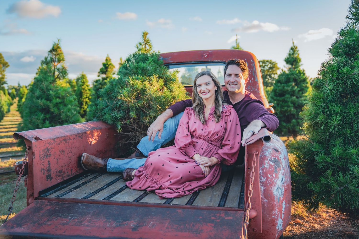 Couple posing in vintage red truck among evergreen trees at sunset.