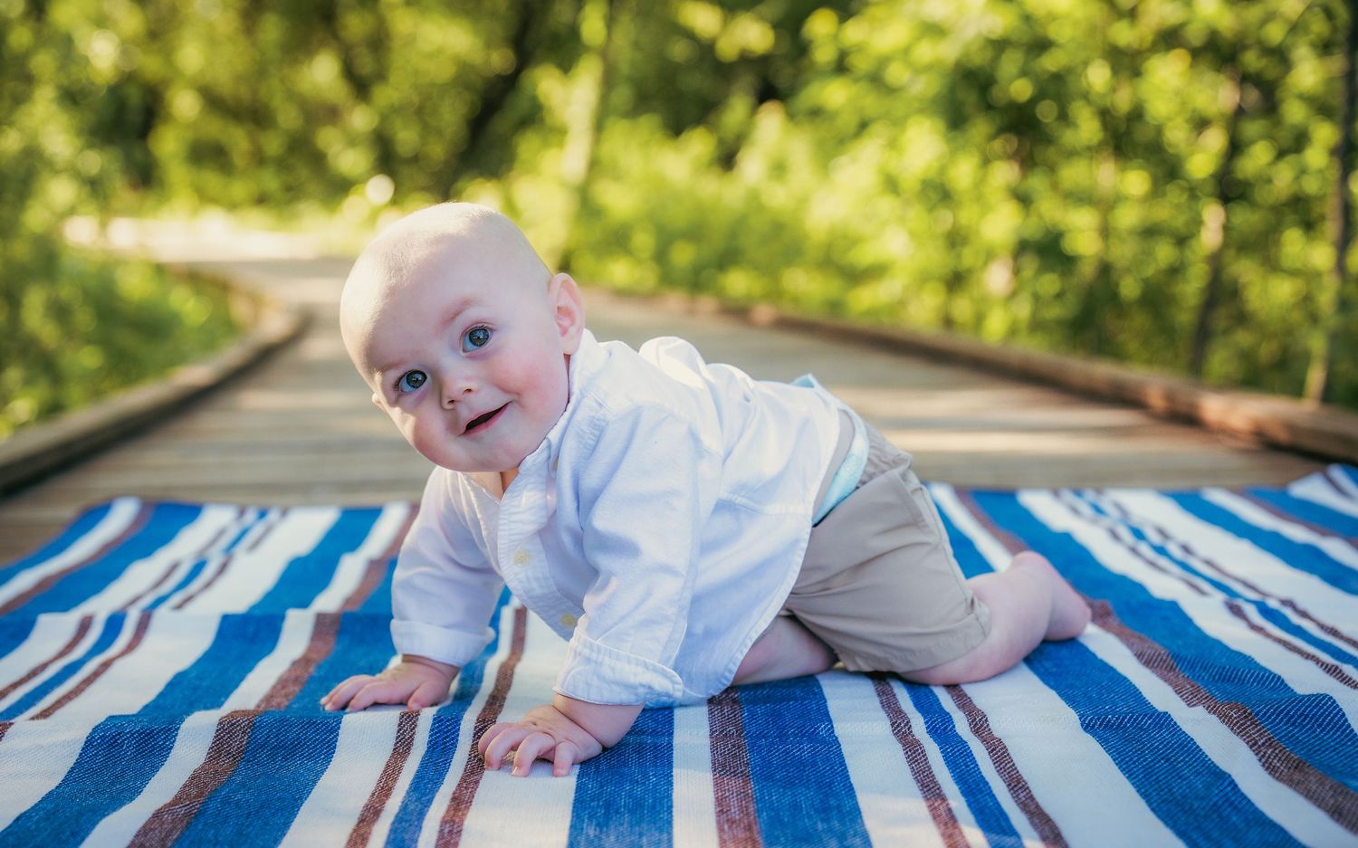 Playful baby crawling on blue and white striped blanket in park setting.