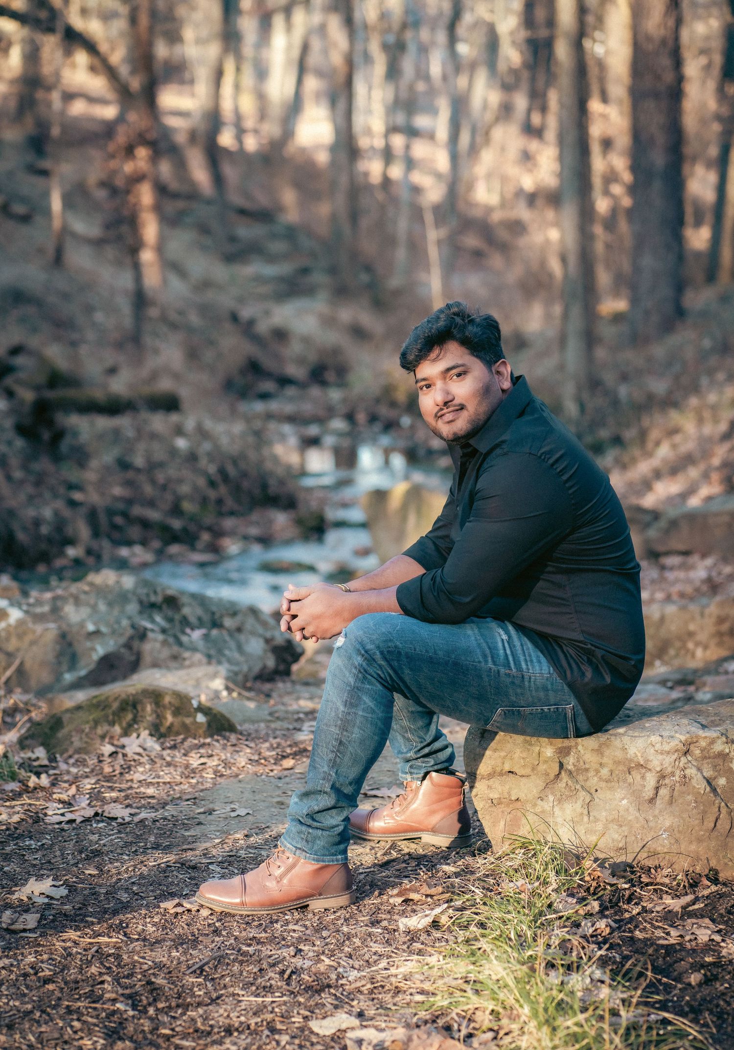 A person in dark clothing sits on rocks in a wooded setting during autumn.