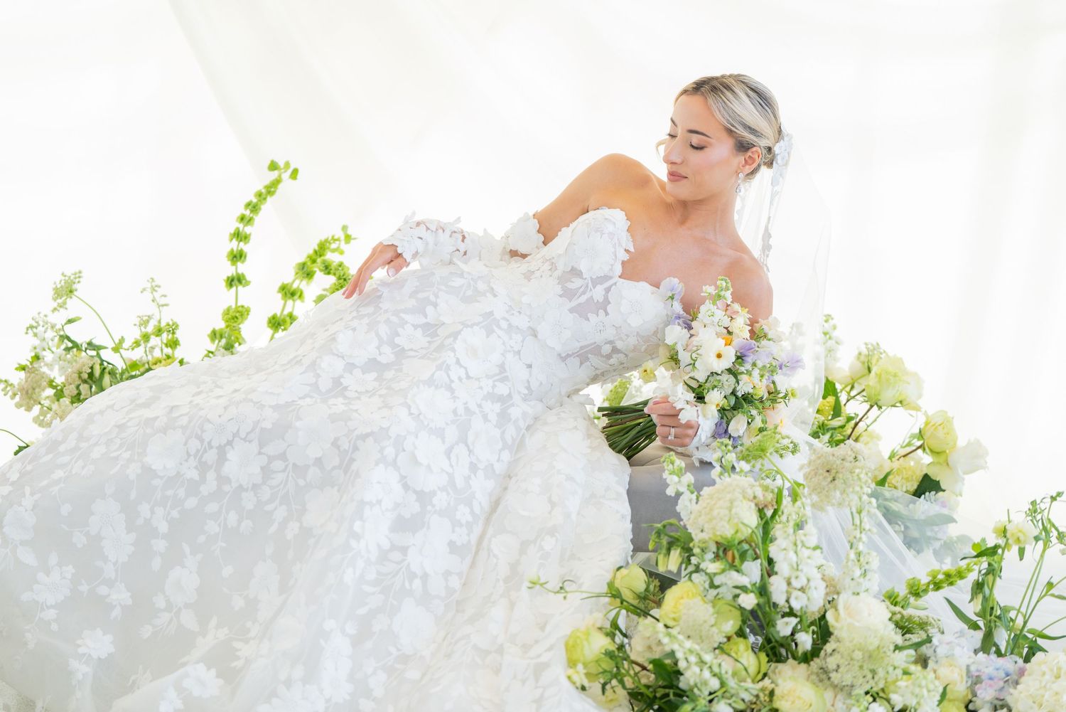 Elegant bride in a voluminous white lace gown poses with floral arrangements against a light backdrop.