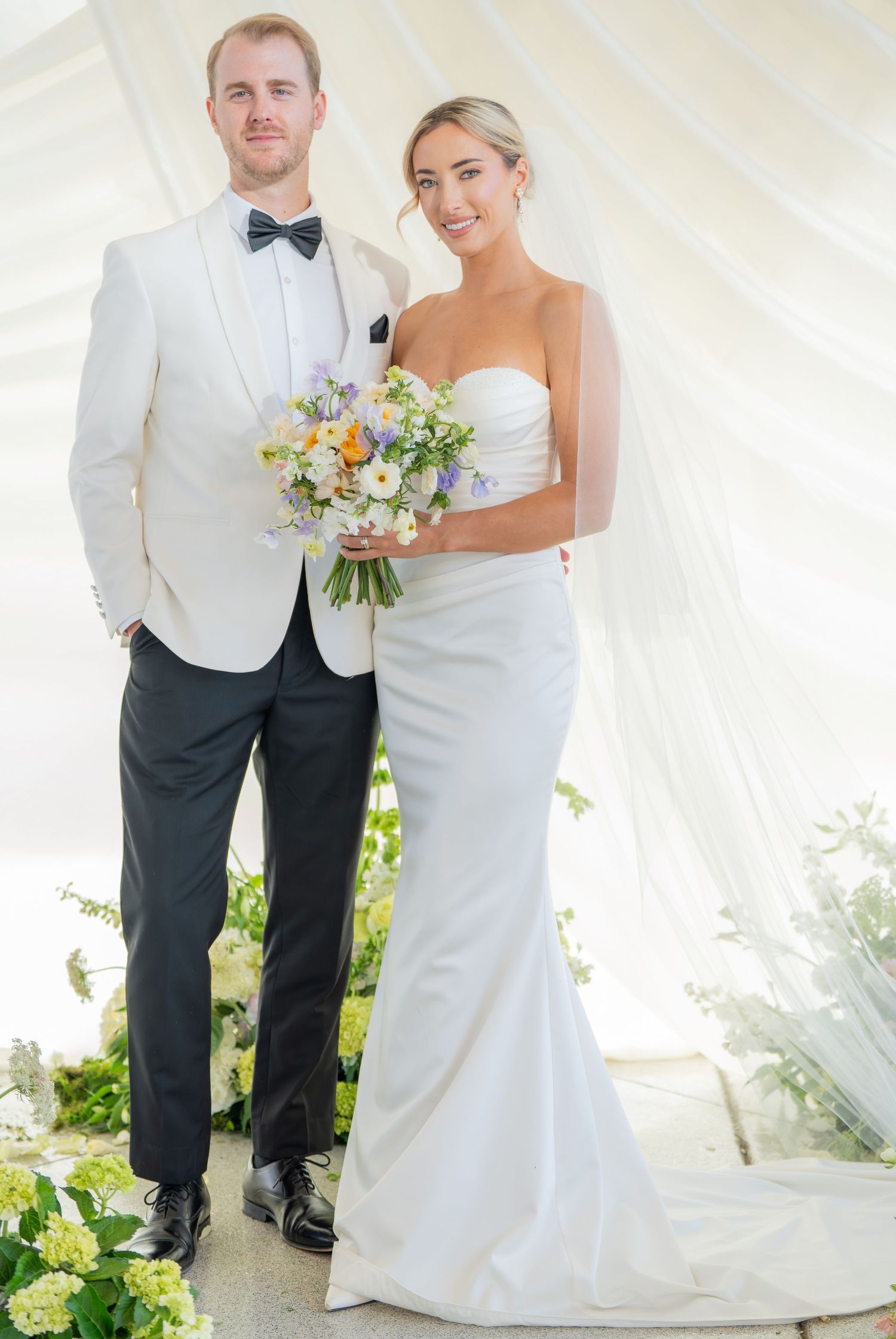Elegant wedding couple poses in a white tent with flowing fabric and floral arrangements.